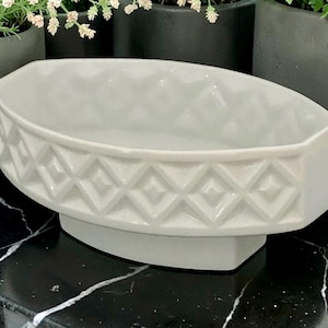 May include: An oval-shaped white ceramic bowl featuring a raised geometric diamond pattern around the exterior sides. The bowl has a smooth, glossy finish and sits on a slightly recessed base, presenting a clean and uniform appearance throughout.