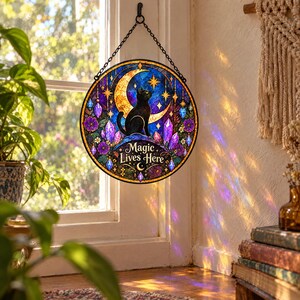 May include: A circular stained glass suncatcher featuring a black cat sitting under a yellow crescent moon and stars. The design includes purple crystals, blue accents, and floral motifs with the text Magic Lives Here, suspended by a black metal chain.