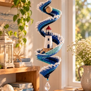 May include: A hanging crochet mobile featuring a central white and blue lighthouse surrounded by a spiraling wave pattern in shades of dark blue, light blue, and white. The structure includes a tan base, a small sphere, and a clear crystal pendant.