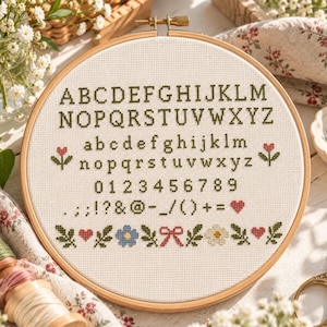 May include: A circular wooden embroidery hoop containing white Aida fabric with a cross-stitched alphabet in uppercase and lowercase letters, numbers zero through nine, various punctuation marks, and a bottom border of flowers, hearts, and a bow.