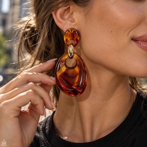 May include: A dangling earring featuring a circular top piece and a larger, irregular oval-shaped bottom piece, both crafted from translucent amber-colored resin with a marbled pattern, connected by a polished gold-tone metal ring link.