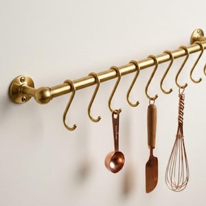 Puede incluir: Un riel de cocina de latón montado en la pared con una barra horizontal, dos soportes circulares y diez ganchos en forma de S extraíbles. Tres utensilios de cocina de tono cobre, incluyendo una cuchara medidora, una espátula y un batidor, cuelgan.