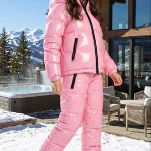Könnte beinhalten: Eine glänzende rosa Daunenjacke mit Kapuze und dazu passender rosa Daunenhose. Die Jacke hat einen schwarzen Frontreißverschluss und schwarze Reißverschlusstaschen. Die Hose hat schwarze Akzente an den Taschen und wird mit weißen Turnschuhen getragen.