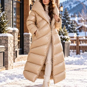 May include: A long, beige, shiny puffer coat with a high collar and a front zipper closure. The coat features horizontal quilted stitching throughout the length, long sleeves, and a full-length silhouette that extends down to the lower legs.