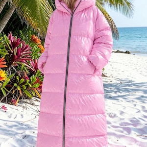 Peut inclure: Un long manteau doudoune rose clair doté d'un col montant, d'une capuche et d'une fermeture éclair noire sur toute la longueur. Le vêtement présente une construction matelassée à bandes horizontales, une finition brillante et deux poches latérales.