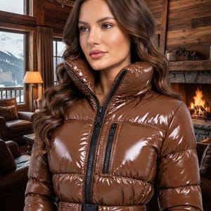 May include: A glossy brown puffer jacket featuring a high collar, a black front zipper, a vertical zippered chest pocket, and a matching belt with a black plastic buckle. The jacket has a quilted design with horizontal stitching throughout.