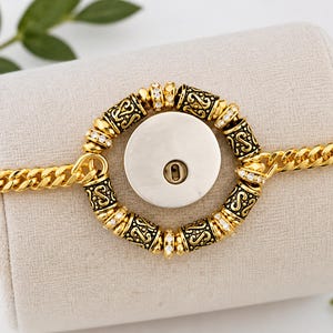 May include: A gold-toned chain bracelet featuring a central circular charm. The charm consists of a smooth, silver-colored disc with a central slot, surrounded by alternating ornate, dark-etched gold beads and small, clear, sparkling stones.