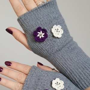 May include: Gray fingerless gloves adorned with crocheted flower embellishments. One glove features a purple flower, the other a white flower. The gloves are made of a soft, knitted material, perfect for cooler weather.