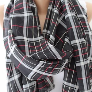 May include: A black scarf with a plaid pattern in white, gray, and red. The scarf is draped around a person's neck, showcasing the fabric's lightweight and flowing texture. The scarf is a fashion accessory.