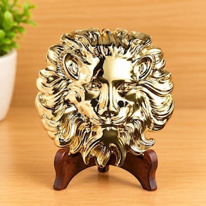 May include: A circular, reflective gold-toned lion head plaque featuring detailed, flowing mane textures and a symmetrical facial expression. The metallic lion sculpture is mounted on a dark brown, curved wooden display stand for upright positioning.