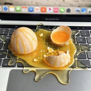 May include: A whole light brown egg and a broken eggshell with a bright yellow yolk are resting on a silver laptop keyboard. The raw egg liquid is spread across the keys and the trackpad area, creating a glossy, viscous puddle on the device.