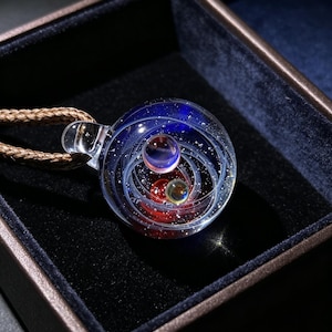 May include: A spherical glass pendant featuring a deep blue and red swirling galaxy design with embedded glittery white specks and two small internal spheres. The pendant is attached to a braided brown cord and rests inside a dark box.