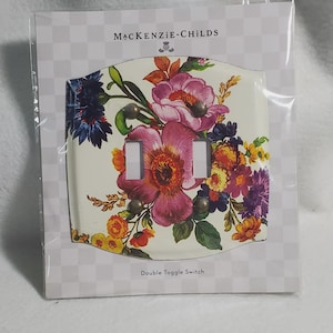 May include: A MacKenzie-Childs double toggle switch plate featuring a vibrant floral pattern with pink, purple, blue, and yellow flowers on a cream background. The rectangular plate has four small metal screws and is sealed in clear plastic packaging.