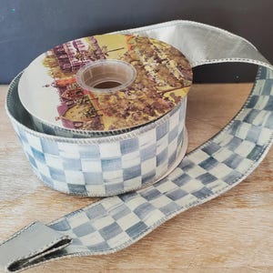 May include: A spool of wired ribbon featuring a blue and white checkered pattern with a painted, brushstroke effect. The ribbon has a metallic silver edge and is wrapped around a cardboard core with a printed illustration of a building.