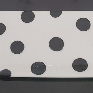 May include: A rectangular ceramic serving tray with a white base and large black polka dots. The tray features two handles, each shaped like a small animal figurine decorated with a black and white checkered pattern and gold-colored accents.