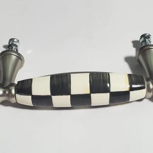 May include: A cabinet pull handle featuring a central cylindrical section with a black and white checkered pattern. The handle has metallic silver-toned ends with a smooth, rounded, and tiered design, including two threaded bolts for mounting.
