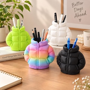 May include: Four puffer jacket-shaped pen holders in neon green, white, black, and a rainbow gradient. Each holder features a high collar, a central zip detail, and ribbed hem, designed to store pens and pencils upright in the neck opening.