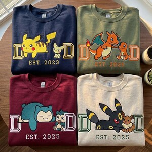 May include: Four crewneck sweatshirts in navy, olive green, maroon, and cream. Each features embroidered Pokémon characters, the letter D on both sides, and text reading EST. 2023 or EST. 2025. The designs include Pikachu, Charizard, Snorlax, and Umbreon.