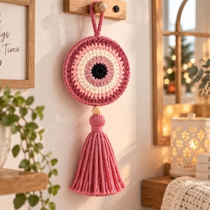 Könnte beinhalten: Ein kreisförmiger Wandbehang aus Häkelarbeit mit einem konzentrischen Augenmuster in Dunkelblau, Weiß und Rosa. Das Stück enthält einen Holzperlenakzent und eine lange, dicke Quaste aus rosa Garn, die an einer Schlaufe aufgehängt ist.