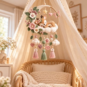 May include: A crochet mobile featuring a crescent moon base with a sleeping fawn, pink flowers, green leaves, a butterfly, and hanging elements including clouds, hearts, small flowers, and tassels in shades of pink, green, and cream.