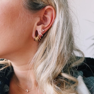 Op de afbeelding: Een set goudkleurige sieraden gedragen in het oor, bestaande uit een cluster van kleine gladde ringetjes in de oorlel, een dun ringetje met ingezette steentjes daarboven en een klein driehoekig knopje bovenaan, met een fijne gouden ketting.