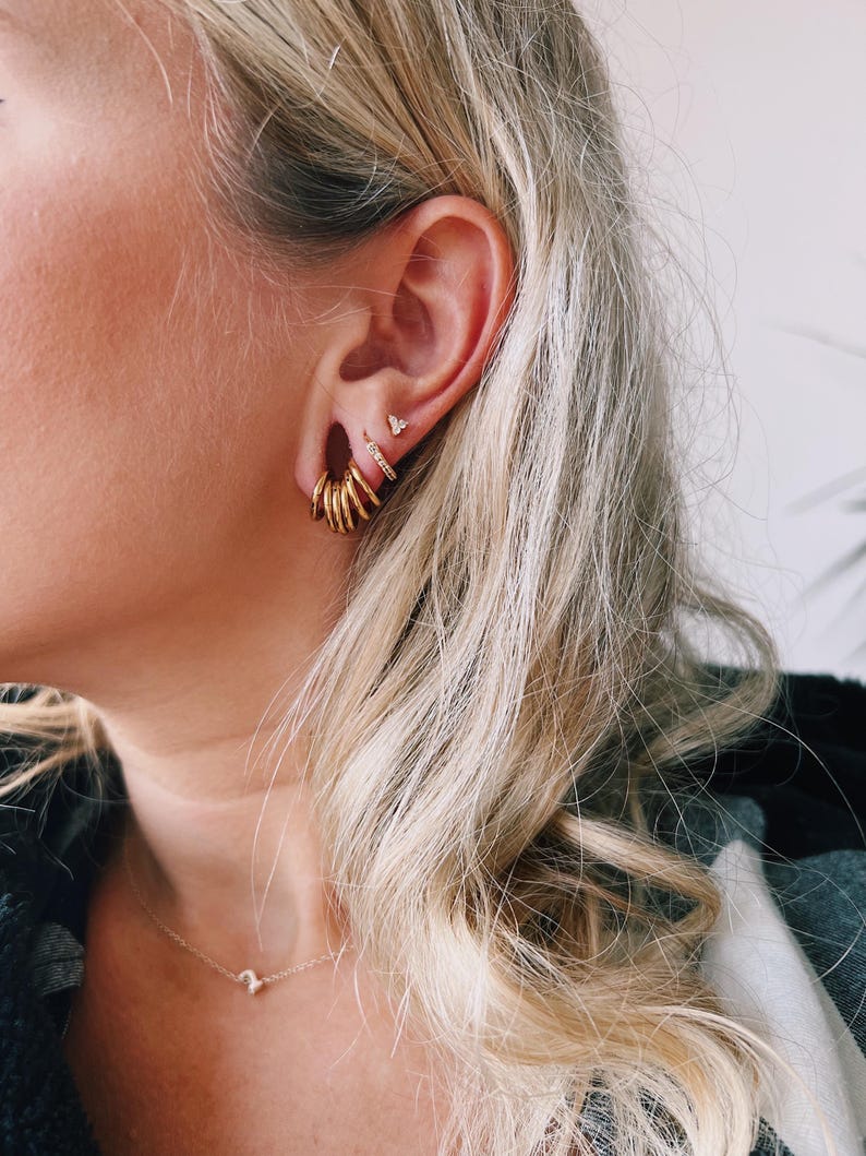 Op de afbeelding: Een set goudkleurige sieraden gedragen in het oor, bestaande uit een cluster van kleine gladde ringetjes in de oorlel, een dun ringetje met ingezette steentjes daarboven en een klein driehoekig knopje bovenaan, met een fijne gouden ketting.