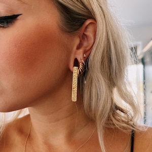 May include: A gold-toned rectangular bar earring with a textured, hammered surface finish. The earring is worn in an earlobe piercing, accompanied by a small gold hoop earring and a tiny triangular stud earring in the upper ear cartilage.