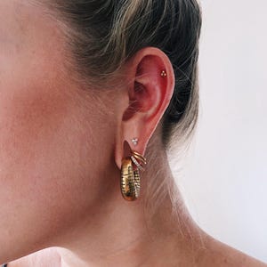 Op de afbeelding: Een goudkleurige oorstack met een grote, getextureerde oorring met een rasterpatroon, twee kleinere gladde gouden ringen, een dunne ring met kristallen, een kleine oorsteker met drie steentjes en een piepkleine gouden driehoekige oorsteker.