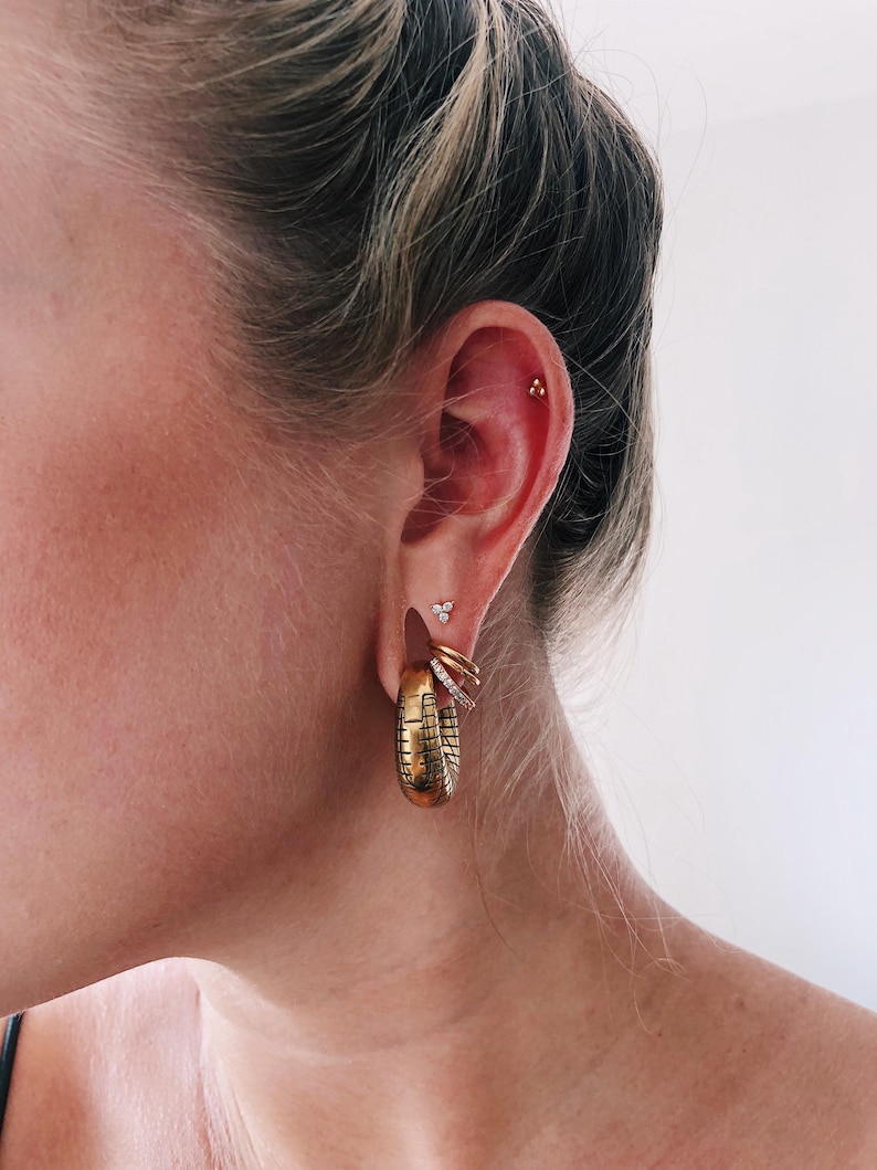 Op de afbeelding: Een goudkleurige oorstack met een grote, getextureerde oorring met een rasterpatroon, twee kleinere gladde gouden ringen, een dunne ring met kristallen, een kleine oorsteker met drie steentjes en een piepkleine gouden driehoekige oorsteker.