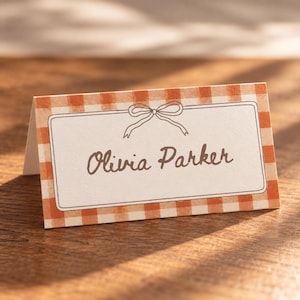 May include: A folded paper place card featuring an orange and white gingham pattern. The center displays a white rectangular area with a hand-drawn brown bow illustration and the name Olivia Parker printed in brown script lettering.