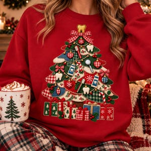 May include: A red sweatshirt featuring a large graphic of a Christmas tree decorated with dental-themed ornaments including teeth, toothbrushes, and toothpaste tubes, with wrapped gift boxes at the base and a white mug with a tree print.