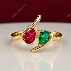 Può includere: Un anello bypass in oro con due pietre preziose taglio a goccia, una rosso vibrante e una verde brillante, incastonate in griffe d'oro lucido. Il design aperto si curva elegantemente attorno al dito, esaltando i colori contrastanti.