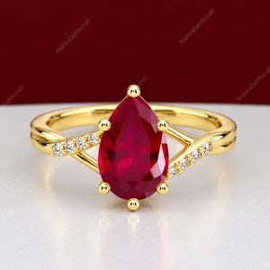 Op de afbeelding: Een gouden ring met een grote peervormige rode edelsteen in het midden, vastgehouden door vijf klauwen. De ringband splitst zich aan beide kanten in twee armen, elk versierd met een rij kleine, heldere, rondgeslepen witte steentjes in het goud.