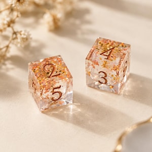May include: Two clear resin dice with embedded gold and copper-colored metallic flakes. The dice are cubic with rounded edges and feature engraved numbers in a dark bronze tone. One die shows the numbers 2 and 5, while the other shows 4 and 3.