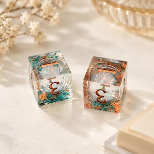 May include: Two square dice made of clear resin, each containing a spherical center with the number 3 visible. The outer cube structure is filled with teal and copper-colored metallic flakes and iridescent glitter, featuring copper-colored numerals.
