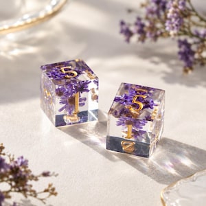 May include: Two clear resin cube dice featuring embedded purple dried flowers and gold-colored numbers. The dice are transparent, showcasing the intricate floral details inside, with sharp edges and a polished finish that reflects light on the surface.