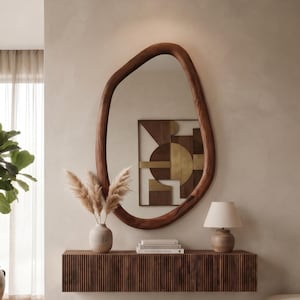 Puede incluir: Una consola flotante montada en la pared con un frente de madera de listones marrón oscuro, rematada con un jarrón de cerámica que contiene hierba de la pampa seca, una pila de libros y una pequeña lámpara de mesa con pantalla beige, bajo un espejo orgánico.