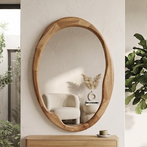 Puede incluir: Un espejo de pared ovalado con un marco grueso de madera clara natural que presenta un borde orgánico e irregular. Montada directamente debajo del espejo hay una consola flotante rectangular de pared hecha de madera clara a juego.
