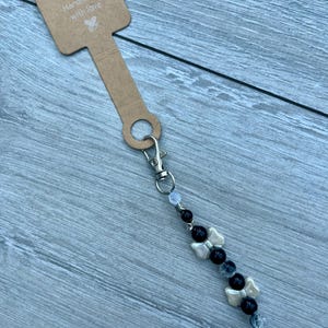 May include: A beaded charm attached to a brown cardboard display tag with the text Handmade with love. The charm features a silver lobster clasp, alternating black round beads, clear faceted beads, and two cream-coloured bow-shaped beads.