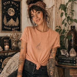 Op de afbeelding: Een perzikkleurig T-shirt met korte mouwen en een kleine, verfijnde lijntekening van een veer of kwastje in het midden van de borst. Het shirt heeft een ronde hals en is gestyled met een geknoopte zoom in de taille, gecombineerd met een donkere jeans.