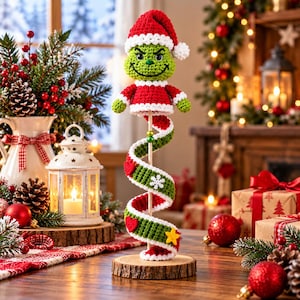 Könnte beinhalten: Eine gehäkelte grüne Figur mit roter und weißer Weihnachtsmütze und Mantel, die auf einem Holzstab sitzt. Eine Spirale aus grün-weißem Häkelstoff, verziert mit roten Herzen, einer weißen Schneeflocke und einem gelben Stern, umwickelt den Stab.