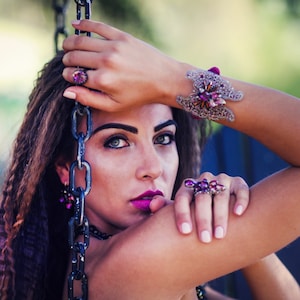 May include: A woman wearing a collection of purple and silver jewelry. The jewelry includes a ring, bracelet, and earrings. The bracelet features a floral design. The woman is holding a chain and looking at the camera.