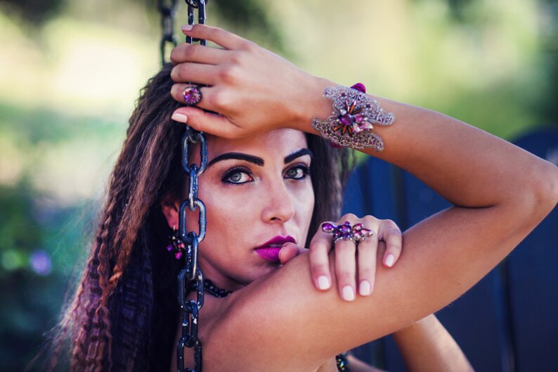 May include: A woman wearing a collection of purple and silver jewelry. The jewelry includes a ring, bracelet, and earrings. The bracelet features a floral design. The woman is holding a chain and looking at the camera.
