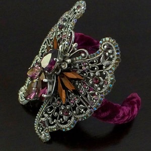 May include: An ornate silver-toned cuff bracelet with a butterfly design. The bracelet features intricate filigree, iridescent rhinestones, and large, faceted purple and amber-colored gemstones. It rests on a plush, dark purple velvet form.