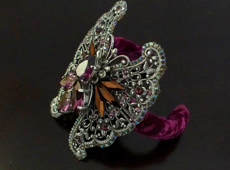 May include: An ornate silver-toned cuff bracelet with a butterfly design. The bracelet features intricate filigree, iridescent rhinestones, and large, faceted purple and amber-colored gemstones. It rests on a plush, dark purple velvet form.