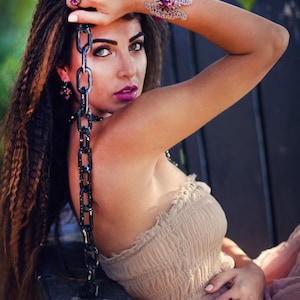 May include: A woman wearing a beige strapless dress and a pink sheer skirt, adorned with jewelry. The jewelry includes a ring, earrings, and a bracelet with purple and pink gemstones. She is seated on a black swing.