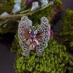 May include: A silver-toned butterfly brooch with intricate filigree detailing. The brooch features a central cluster of purple and pink gemstones, with brown and clear accents. The wings are adorned with smaller purple crystals. The brooch is set against a backdrop of green moss.