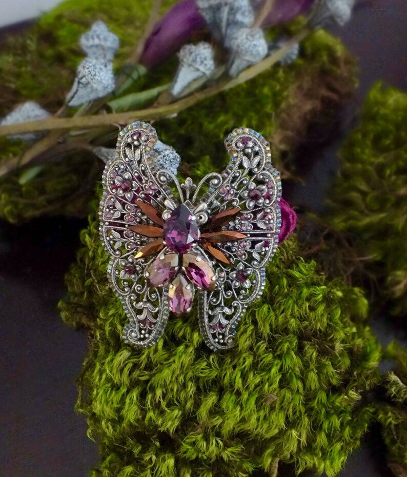 May include: A silver-toned butterfly brooch with intricate filigree detailing. The brooch features a central cluster of purple and pink gemstones, with brown and clear accents. The wings are adorned with smaller purple crystals. The brooch is set against a backdrop of green moss.