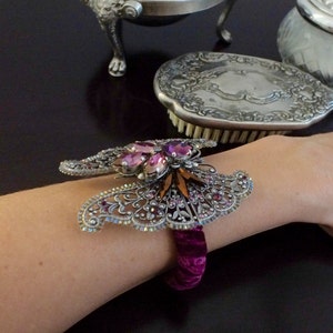 May include: A silver-toned butterfly bracelet with pink and orange gemstones and a deep purple velvet band. The bracelet is worn on a person's wrist, with a dark background.