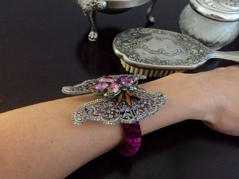May include: A silver-toned butterfly bracelet with pink and orange gemstones and a deep purple velvet band. The bracelet is worn on a person's wrist, with a dark background.